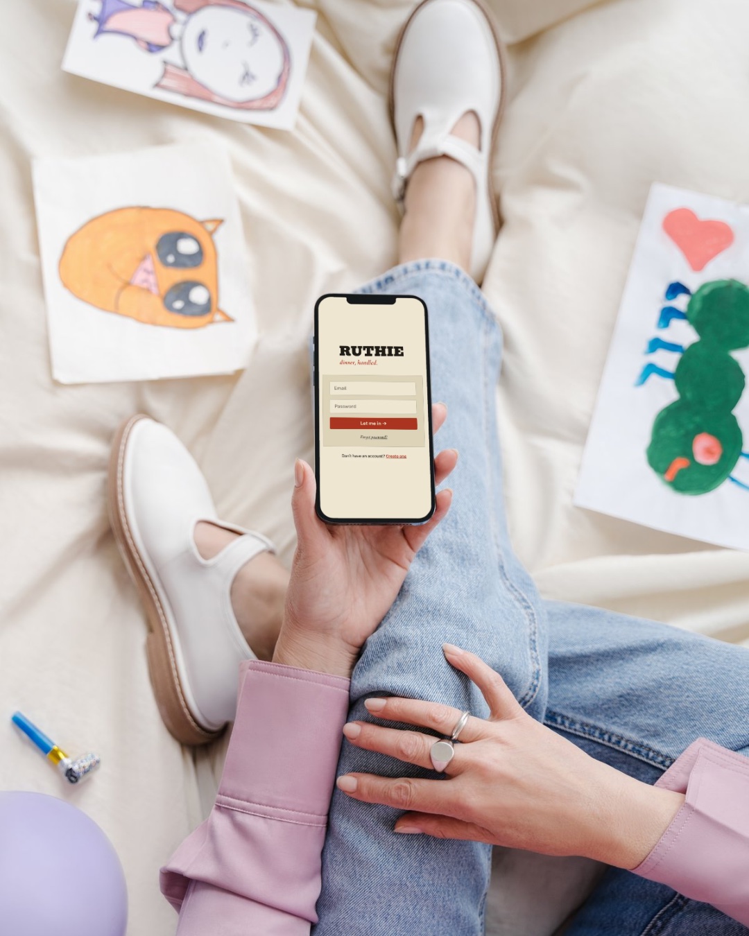 A hand holding an iPhone running the Ruthie app, surrounded by children's crayon drawings, sneakers, and jeans on a soft cream bedspread